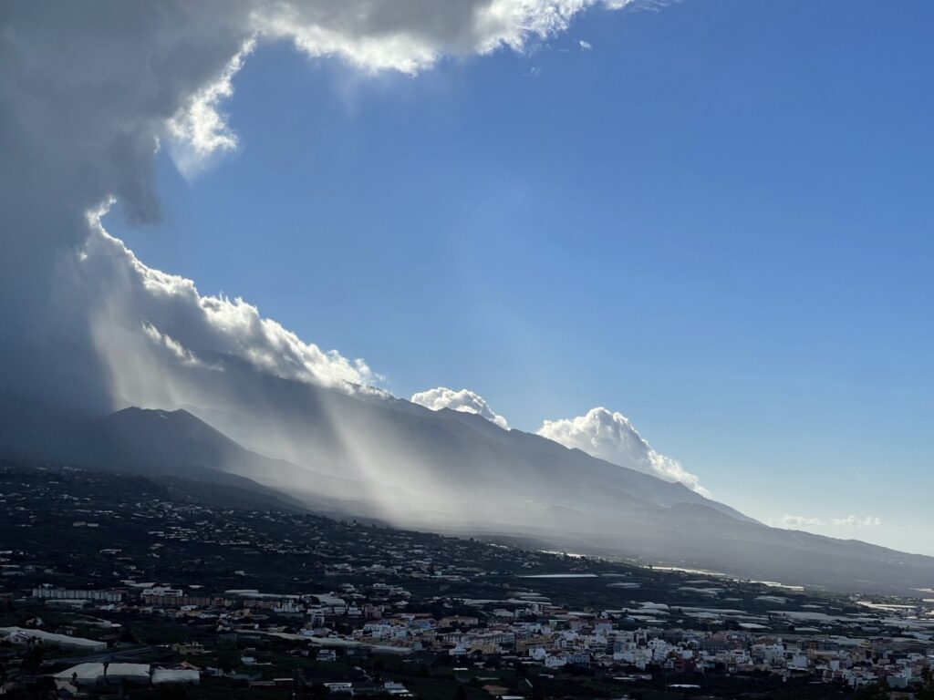 Panorama über das Aridane-Tal mit Ort und Bananenfeldern, dahinter Bergketten mit Regenschauern und Wolken vor blauem Himmel