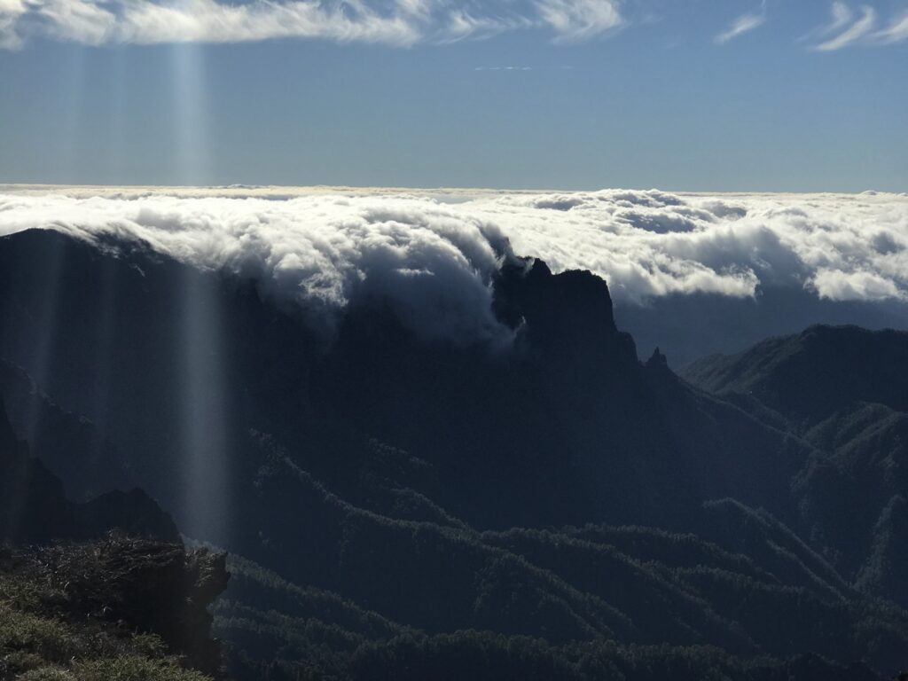 Weißes Wolkenmeer strömt über dunkle Bergkämme der Caldera de Taburiente, darüber tiefblauer Himmel und Sonnenlichtstrahl