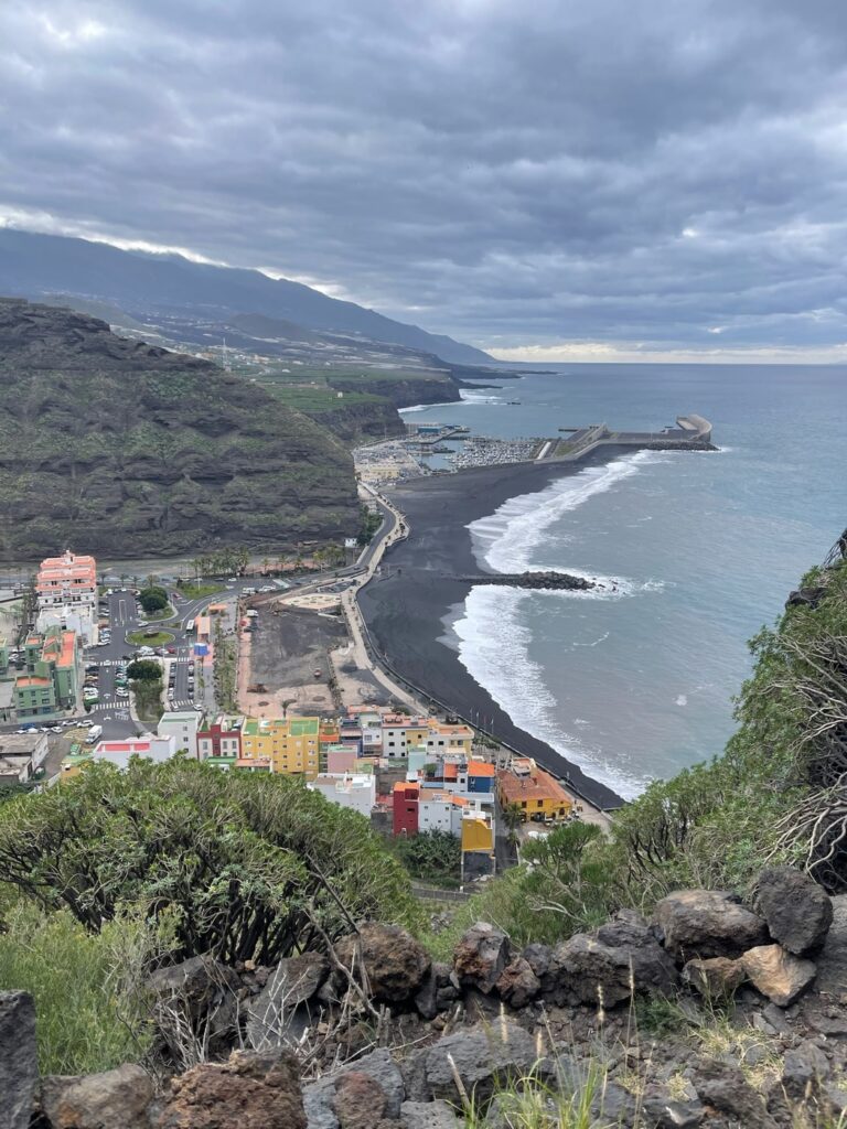 Erhoehter Panoramablick vom Mirador El Time auf den Kuestenort Puerto de Tazacorte mit schwarzem Strand und Hafen unter bewoelktem Himmel