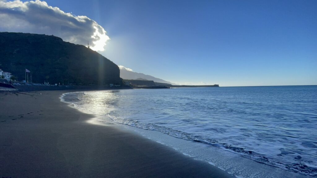 Schwarzer Sandstrand von Puerto Naos mit auslaufender Brandung, tief stehender Sonne hinter dem Berghang und Hafenmole im Hintergrund