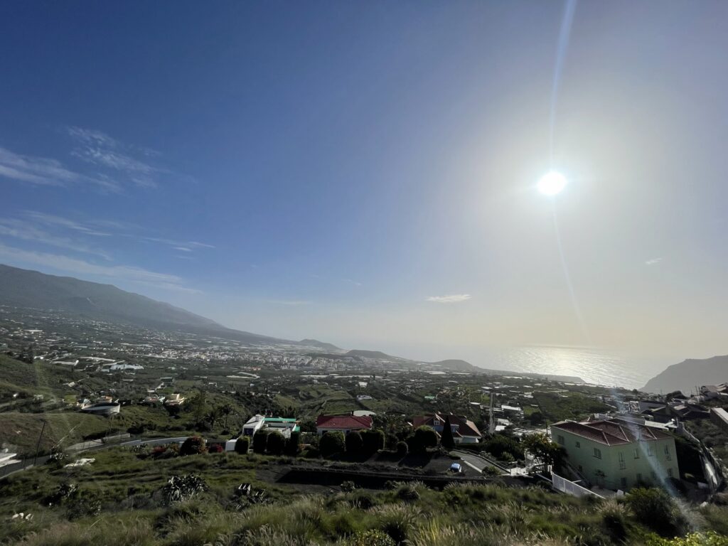 Tagespanorama ueber das Valle de Aridane mit Ortschaft, Feldern, Berghang und Meer unter blauem Himmel mit Sonne
