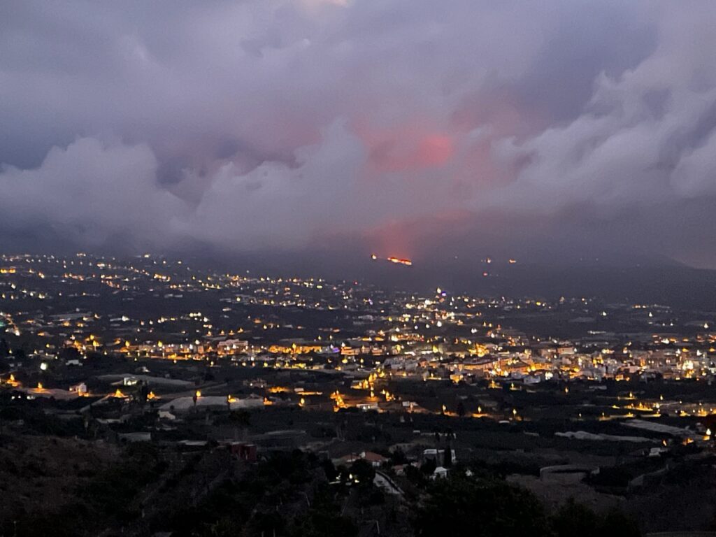 Abendlicher Blick über das beleuchtete Aridane-Tal auf La Palma mit rot glühendem Vulkanausbruch am bewölkten Horizont