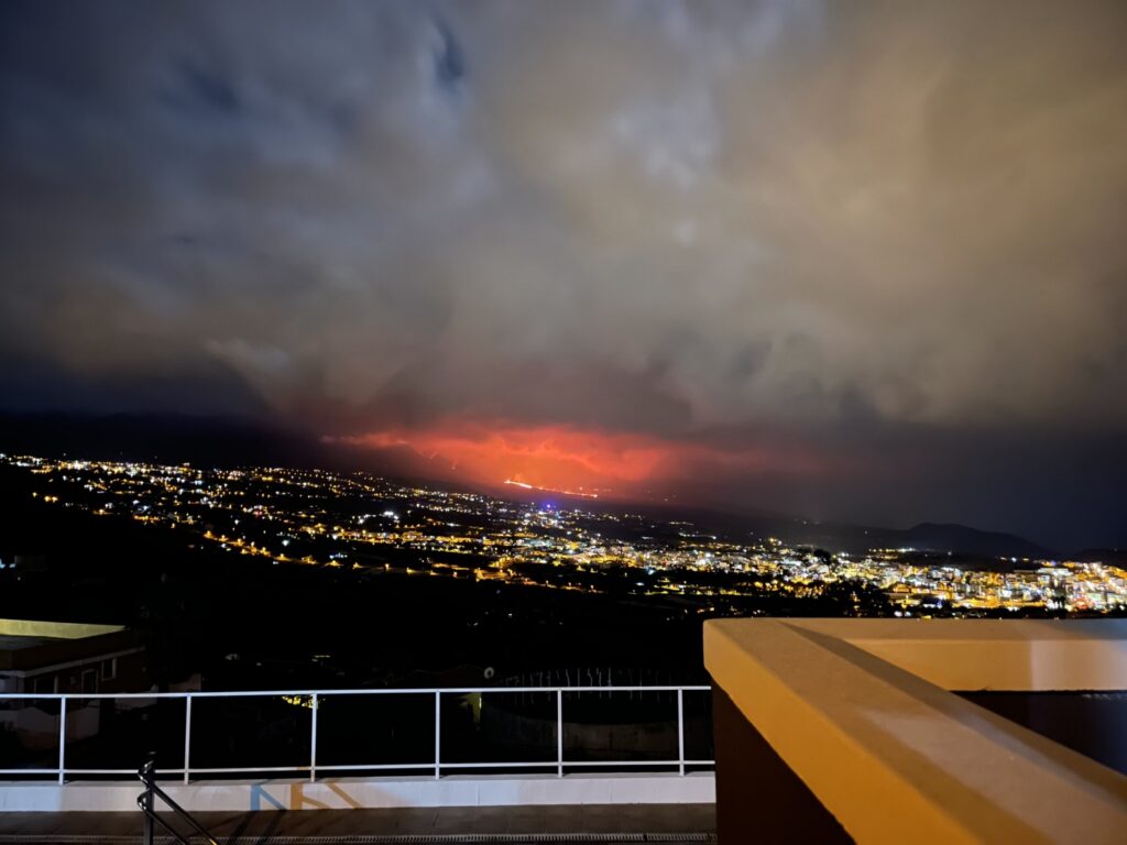 Nachtaufnahme einer Dachterrasse mit Geländer, den Lichtern des Aridane-Tals und orangem Vulkanglühen am Horizont unter dunklen Wolken